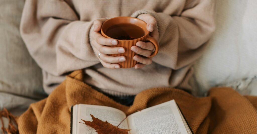 Person holding a warm drink above an open book and blanket, symbolizing the reflective tone of All the Colors of the Dark by Chris Whitaker.