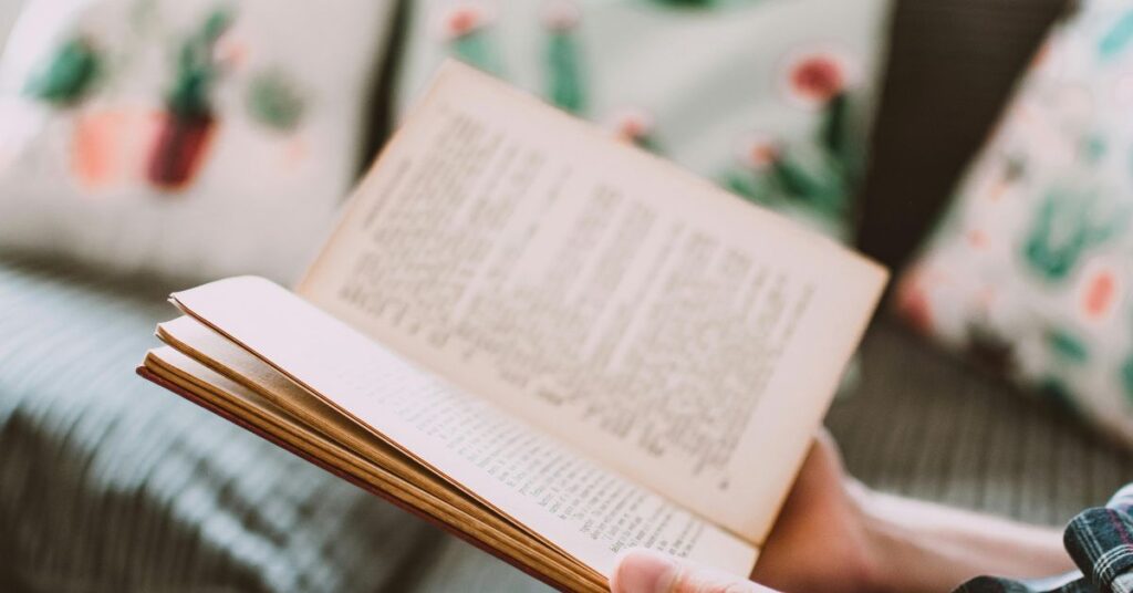 Person holding an open book while reading on a couch with soft light and patterned pillows, representing a calm reading moment.